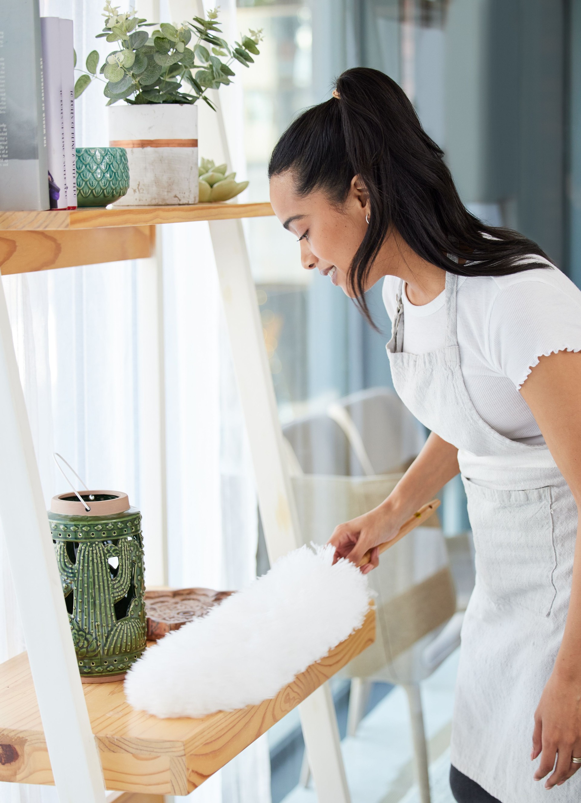 Shot of an attractive young woman standing alone in her home and dusting her shelves
